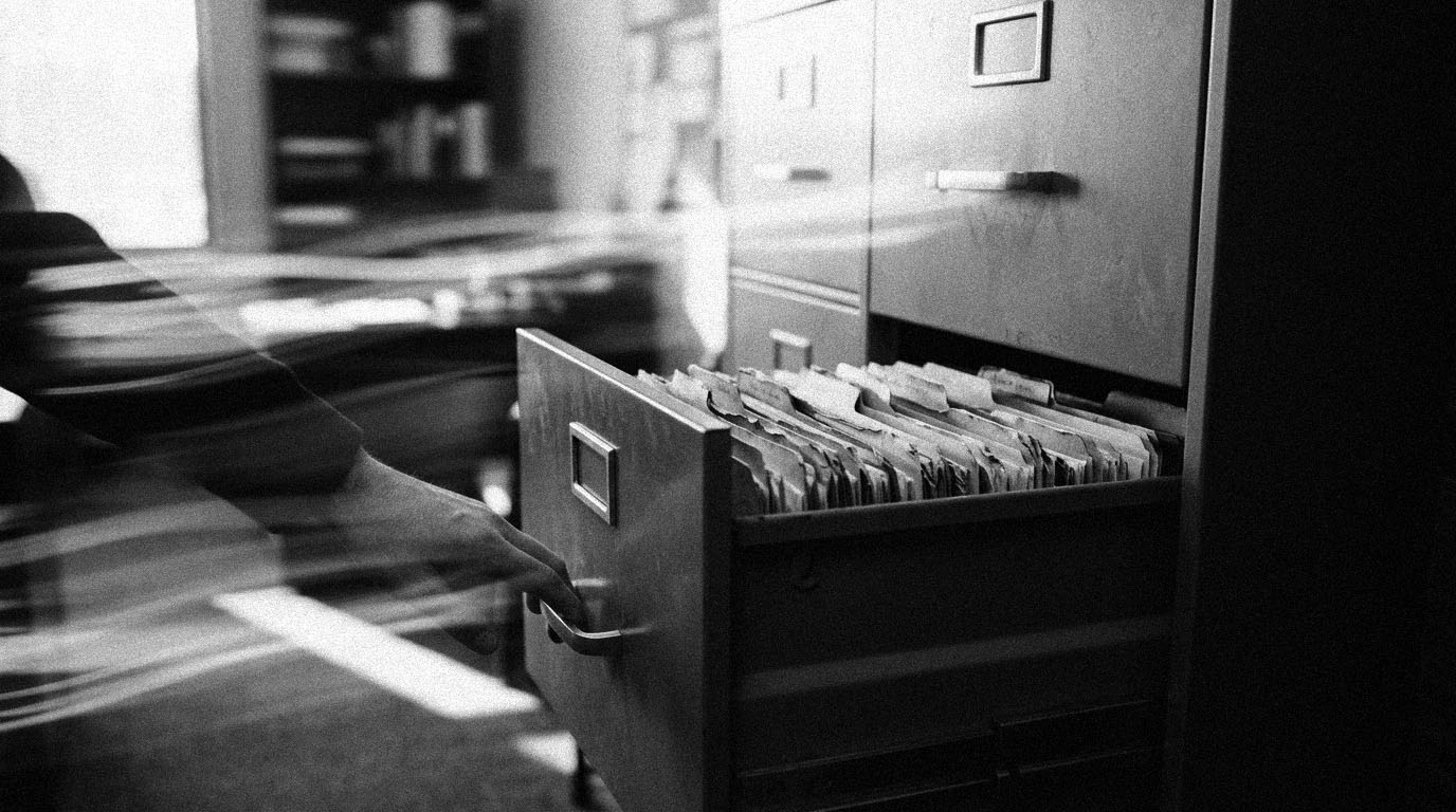 A hand on the handle of an open steel filing drawer of worn manila folders in an office, in black and white.